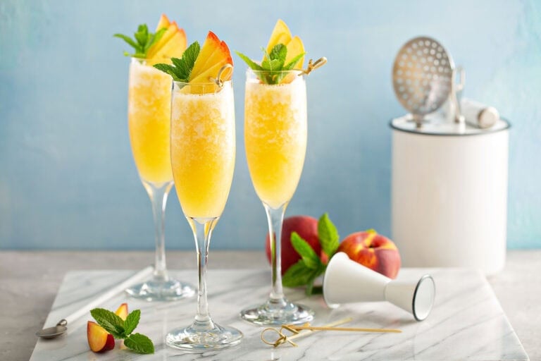 Mimosa bar with three champagne glasses filled with fresh peach mimosas on a marble cutting board