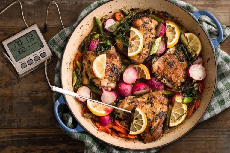 Digital thermometer stuck in chicken thigh in enameled cast iron dish on distressed wood table